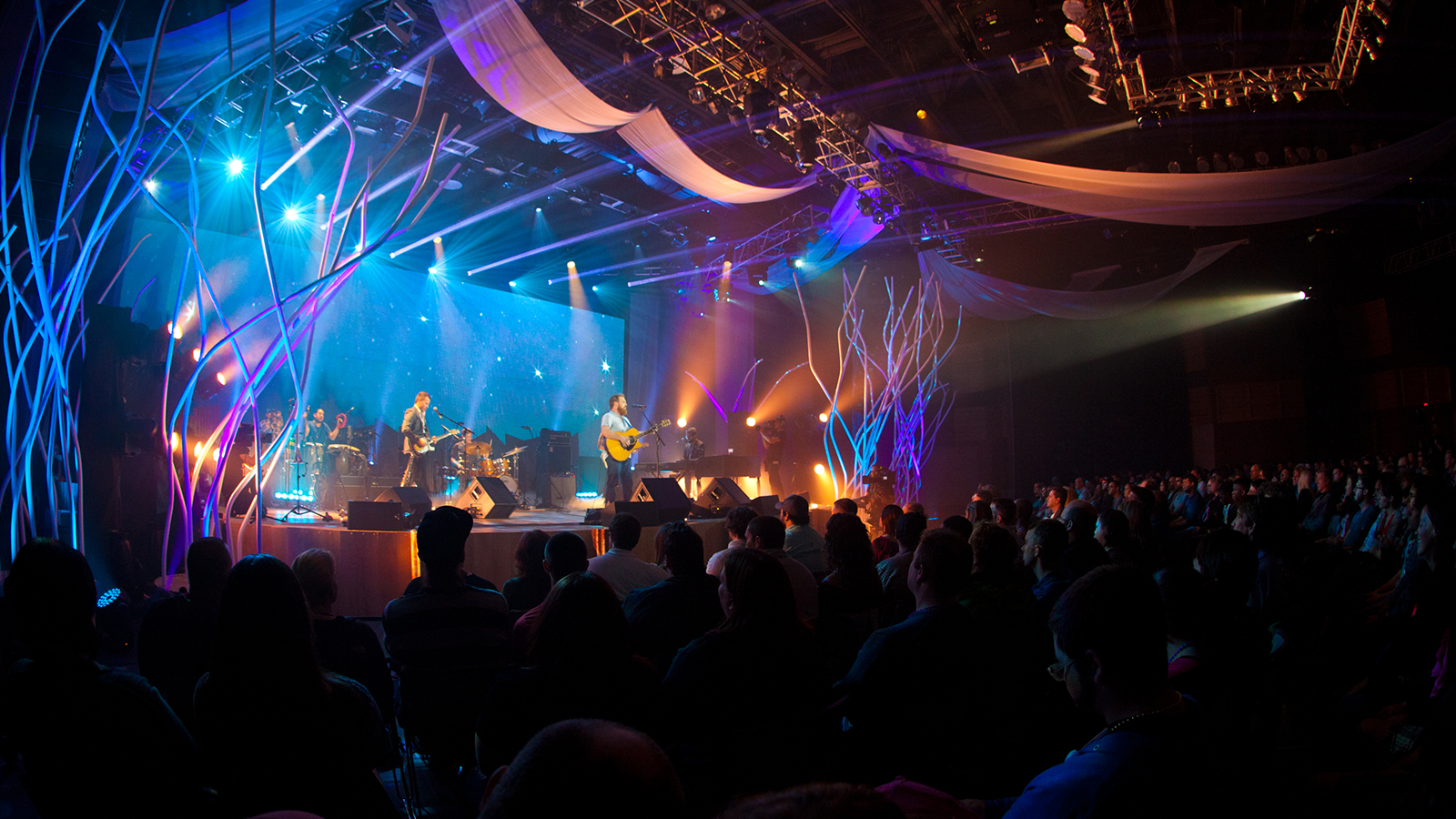 A band plays music in a large, dark auditorium with several bright blue and purple lights pointing at the stage.