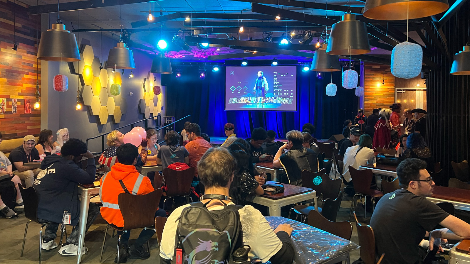 A crowd of students sit at tables in a large room. A large screen displays a video game and decorative paper lanterns hang around the room.
