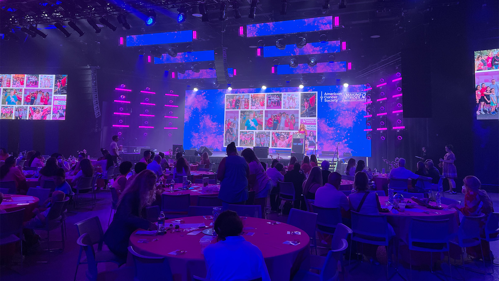Groups of people sit around circular tables in a large room with pink and blue lights. A large screen displays photos and the American Cancer Society Making Strides Against Breast Cancer logo