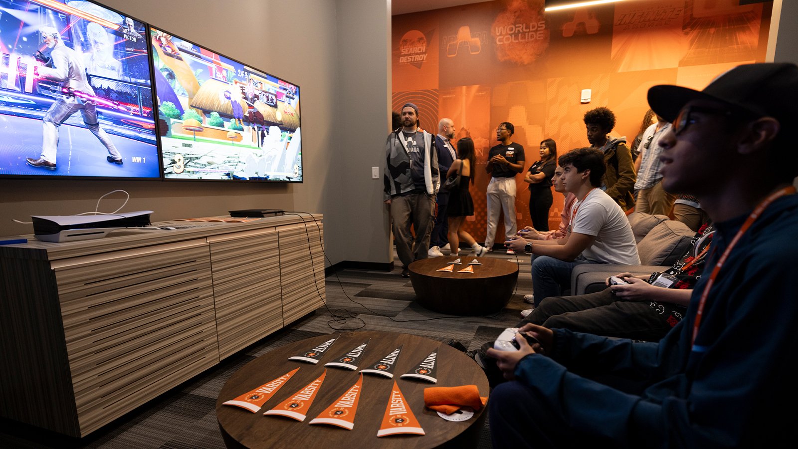 Gamers compete in a lounge with dual screens, a PlayStation 5, and Armada-branded decor, while a crowd watches in the background.