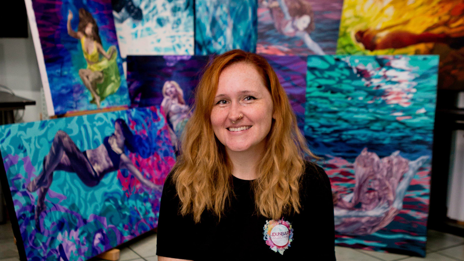 Jessica sits in front of several of her colorful paintings. She is wearing a black t-shirt and is smiling.