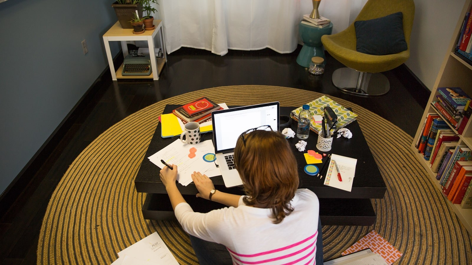 A student sits cross-legged on the floor in front of a coffee table with a laptop, papers, notebooks, and pens.