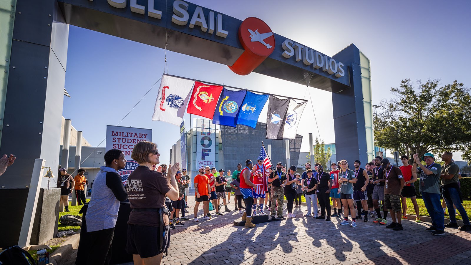 Three 5K winners stand on a podium with medals around their necks under the Full Sail Studios archway. Other competitors stand near them and applaud.