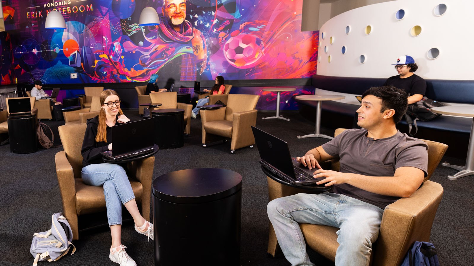 Students study and relax with laptops in Full Sail’s 3B Art Building Lobby, featuring colorful mural art and lounge seating.