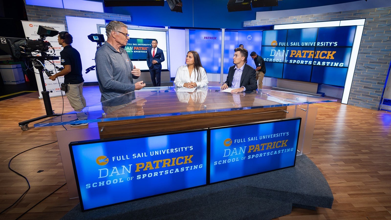 Sportscaster Dan Patrick standing at a news desk speaking with two students at the Full Sail University Dan Patrick School of Sportscasting soundstage.