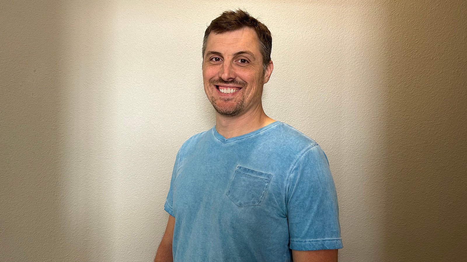 James Lane stands in front of a beige wall and smiles at the camera. He is wearing a light blue v-neck t-shirt.