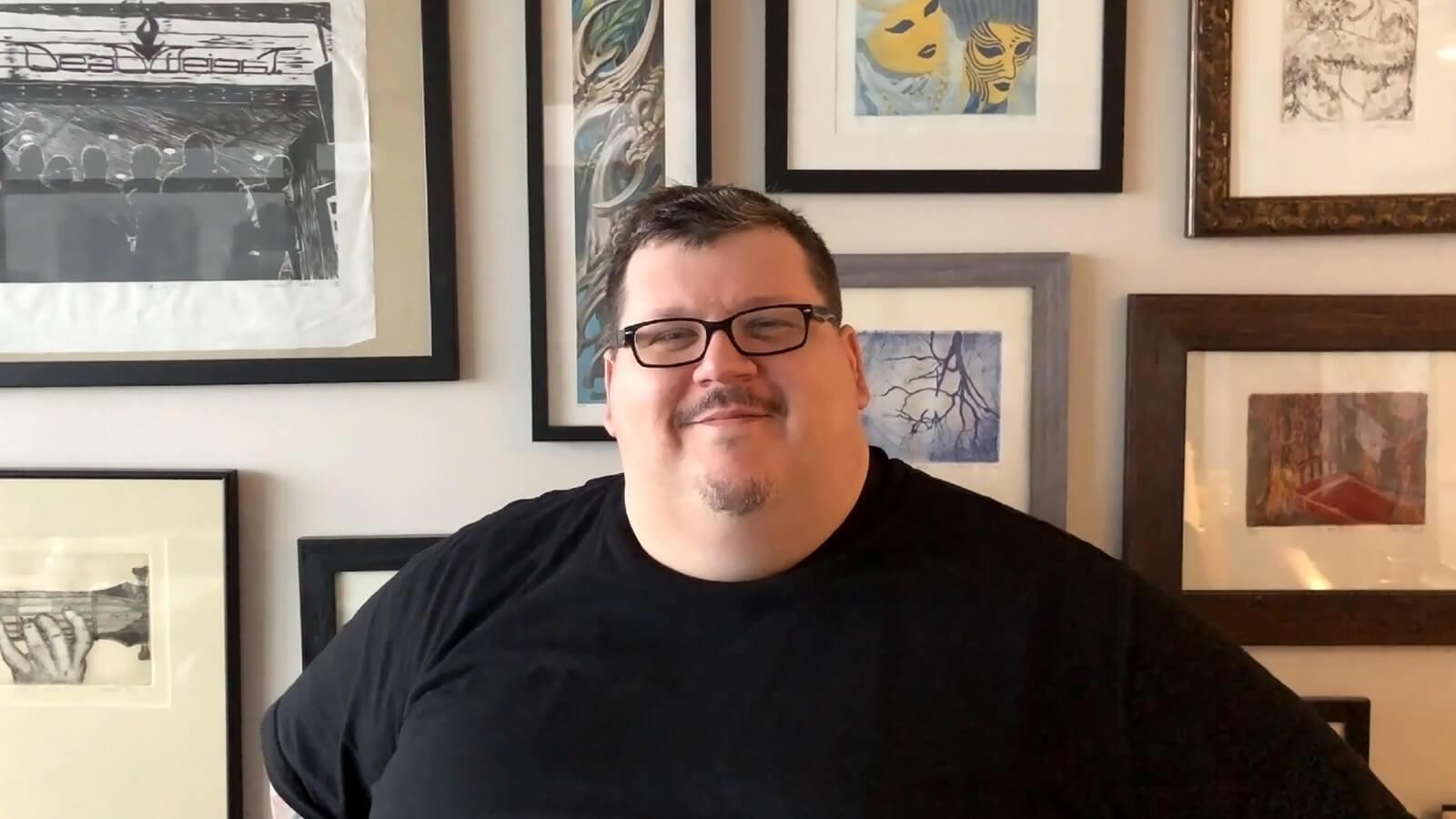 A smiling man wearing glasses and a black t-shirt sits in front of a wall covered with framed paintings.