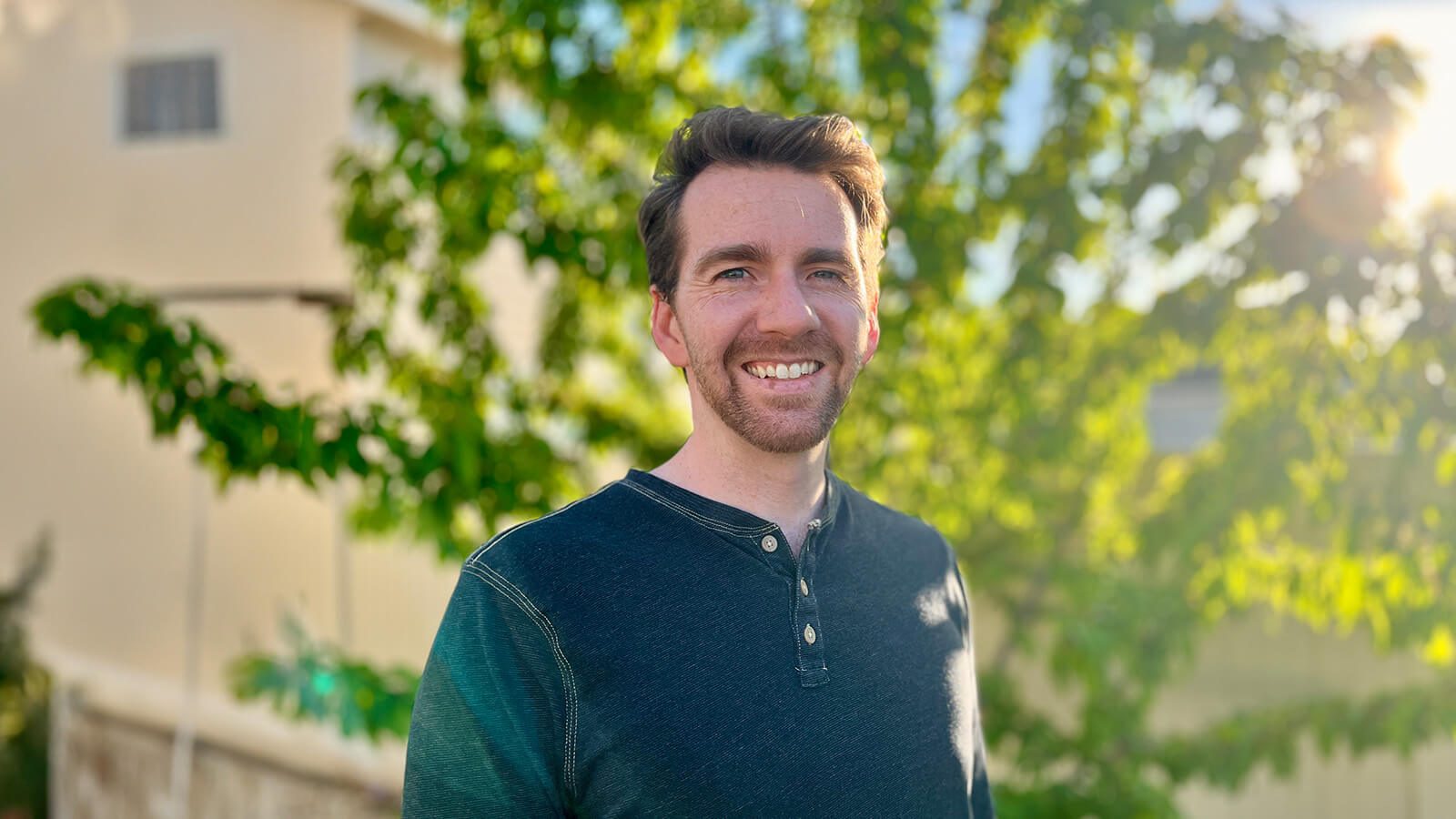 Lee Anderson stands outdoors in front of a tree and smiles at the camera. He has red hair and a beard, and he’s wearing a teal henley.