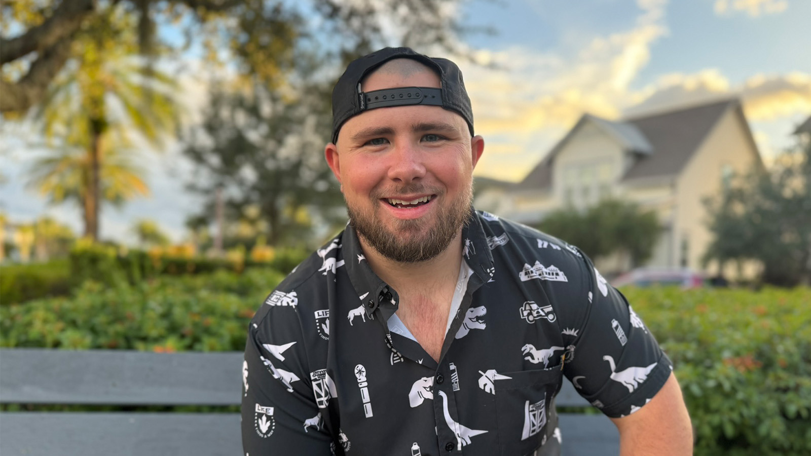 Tyler Yates sits on a bench in an outdoor park and smiles. He’s wearing a black and white collared shirt with dinosaurs on it and a backwards cap.