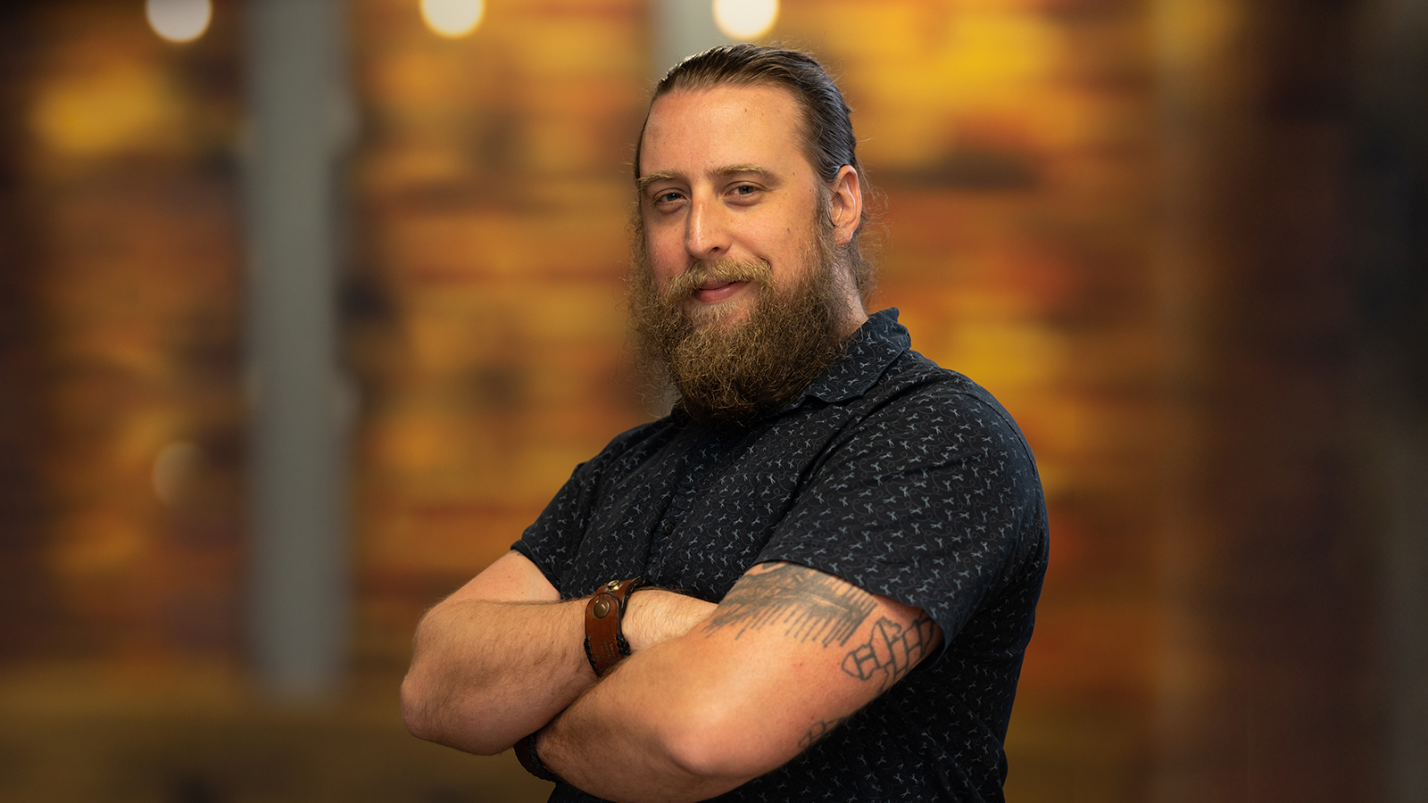 Kyle is standing with his arms crossed, smiling gently. He is wearing a short sleeve blue shirt and his hair is in a bun.