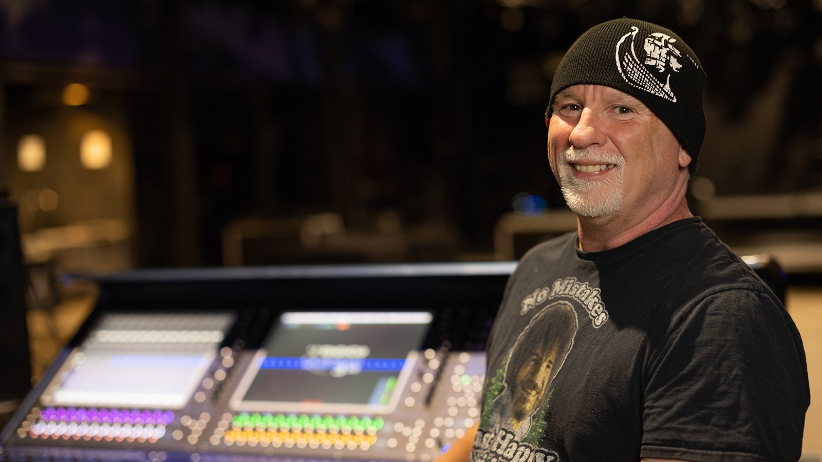 Gregg in a sound studio wearing a black beanie and graphic t-shirt. He is smiling.