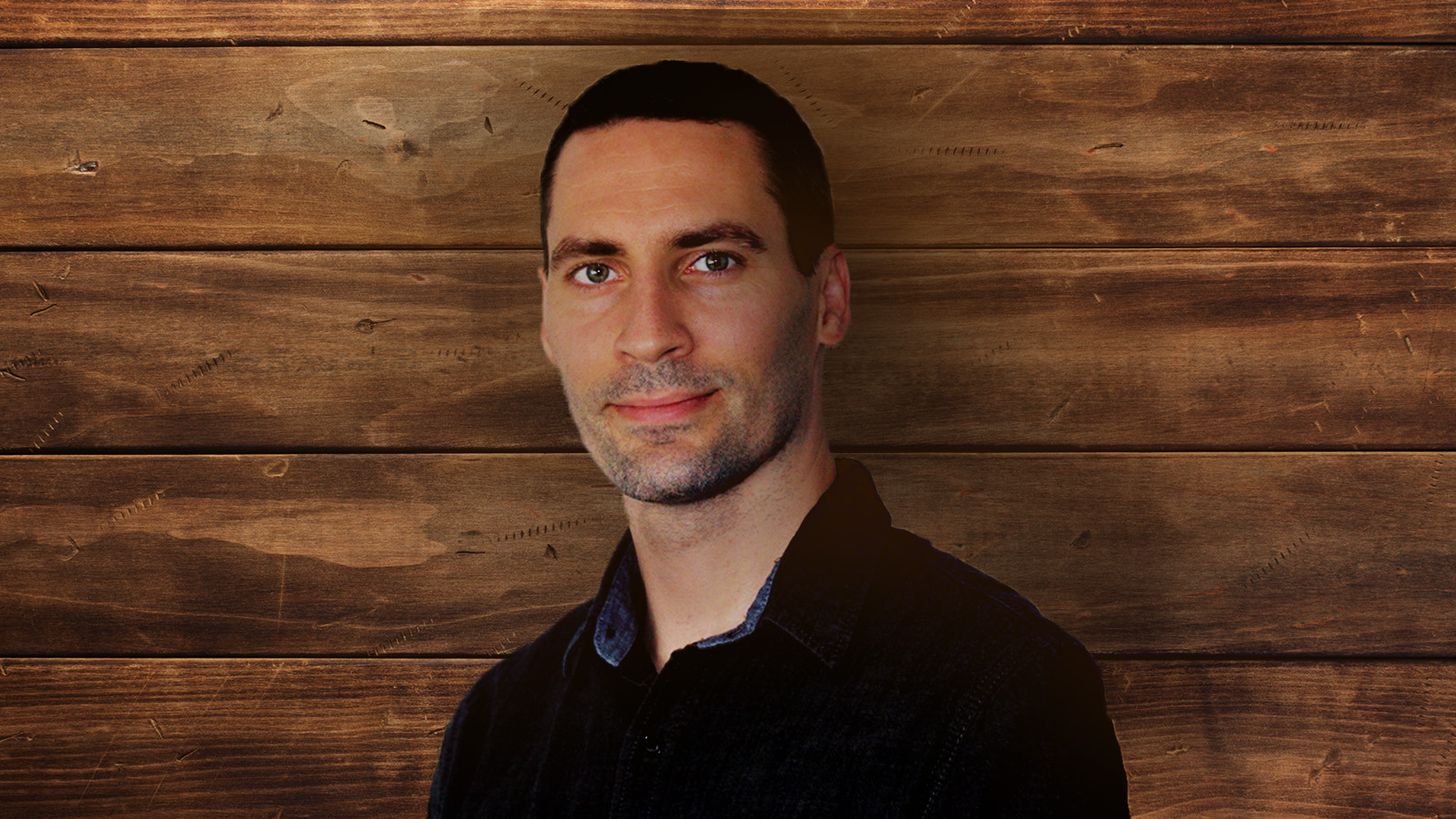 Justin Hosie standing in front of a paneled wood background. He is wearing a dark denim shirt and gently smiling.