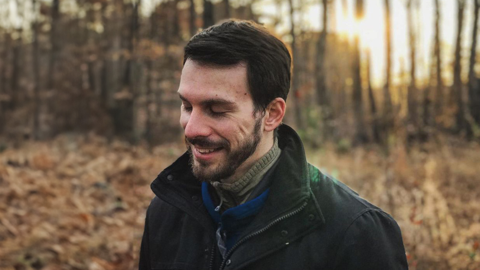 Mikey Zoppo stands in a forest with sunlight poking through the trees behind him. He looks down and smiles, and he is wearing a dark jacket.
