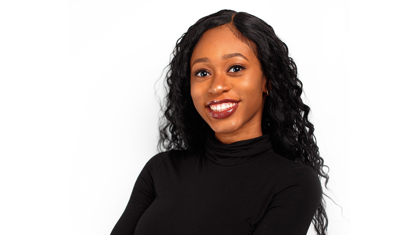 A young woman with long black curly hair, smiling with her arms crossed over her midsection while wearing a long-sleeve black turtle neck against a white backdrop.