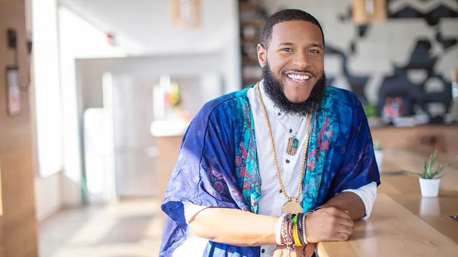 Rico Robinson stands in a café. He is wearing a blue-patterned wrap over a white button-up shirt. He is smiling.