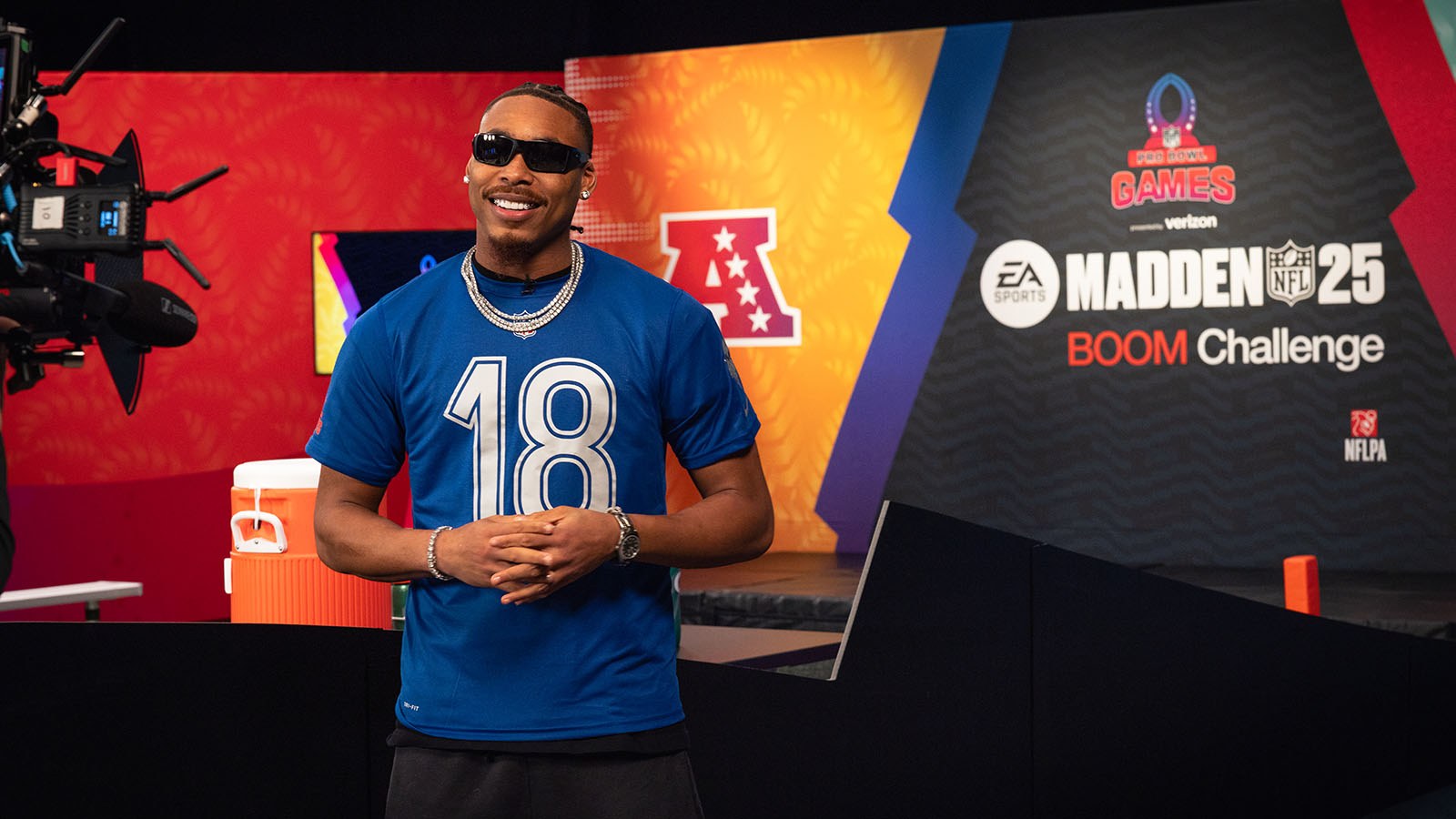 Minnesota Vikings wide receiver Justin Jefferson smiles while wearing a blue NFL-branded t-shirt with the number '18' in a studio set for the Madden NFL 25 Boom Challenge.