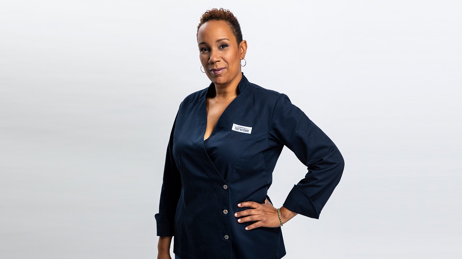 Grad Elle Simone Scott standing and smiling against a white backdrop while wearing a blue double-breasted chef's jacket with an America's Test Kitchen patch on the breast.