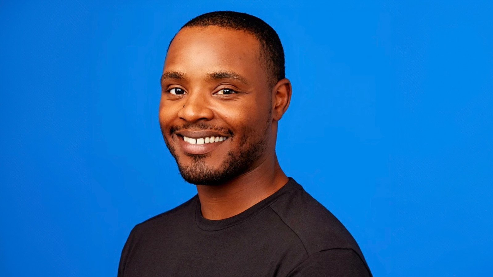 Lamont Johnson smiles at the camera in front of a blue background. He is wearing a black shirt.