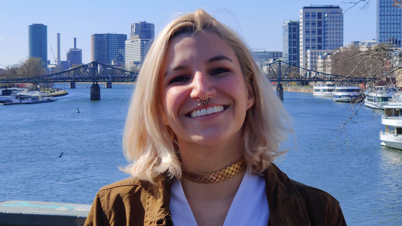 Grad Angela Decastro, a woman with shoulder-length blonde hair wearing a brown jacket and white top, smiles while standing on a bridge overlooking a river, in the distance behind her is a bridge and cityscape.