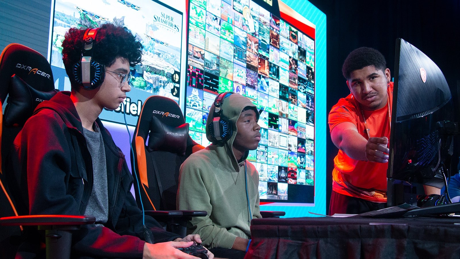 Two Florida high school students seated on stage in front of a gaming monitor with a Full Sail Armada athlete helping them to their right.