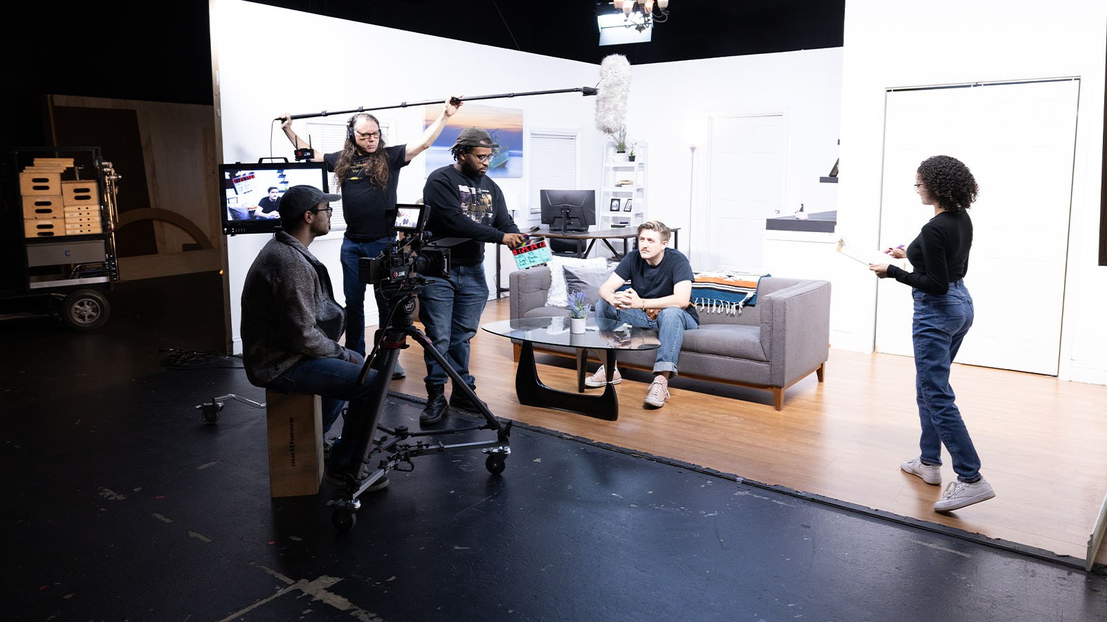 A group of students work together in a living room set on one of Full Sail’s soundstages. They are using a camera and holding microphones and a script.
