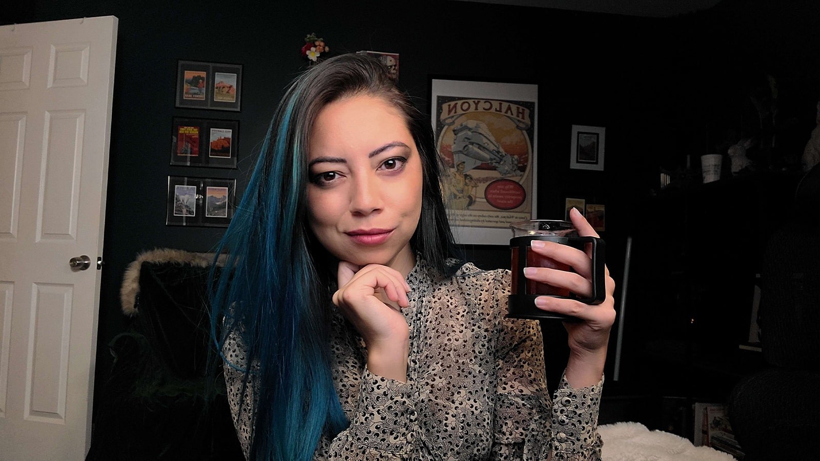 A woman with long brown hair with blue highlights holding a mug while seated in a home office.