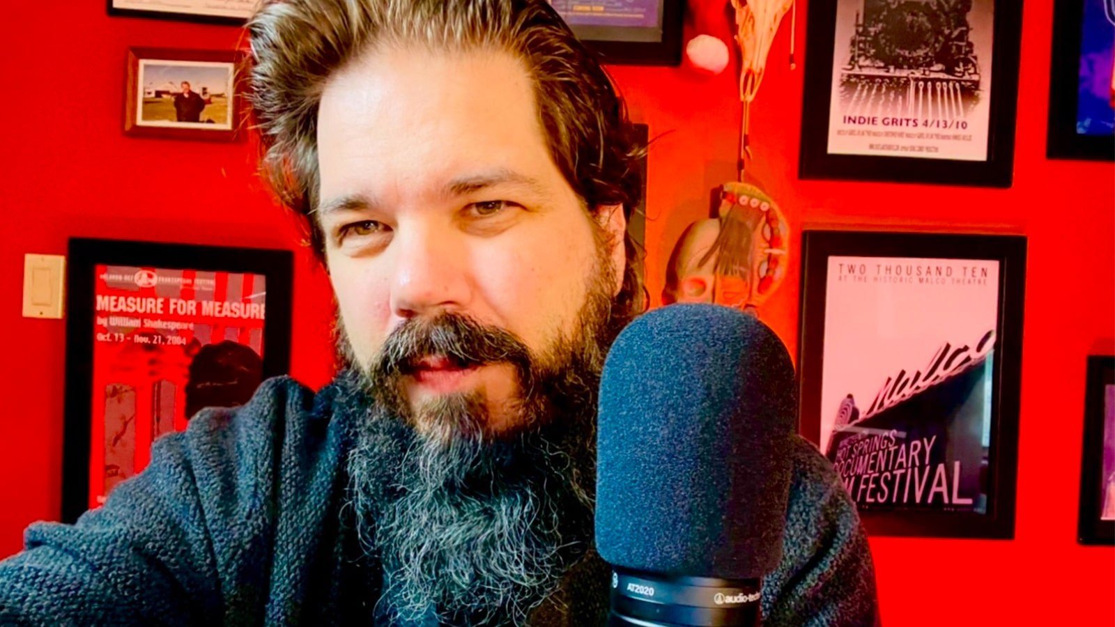 Christopher Ramsey, a man with a long black and grey beard and short hair sitting with a microphone in front of a red wall filled with framed film posters.