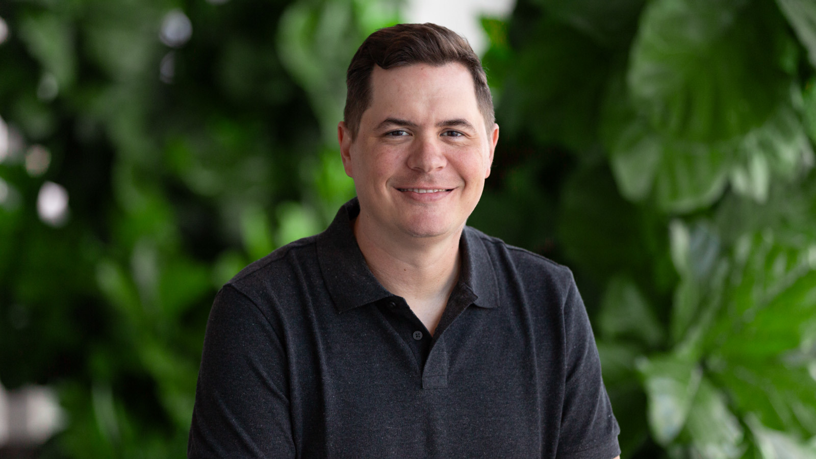 A man with short brown hair is wearing a dark grey polo while smiling against a lush green backdrop.