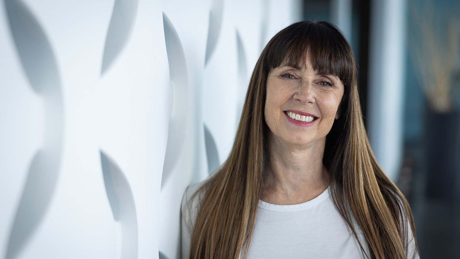 A woman with long brown hair and bangs smiling while standing against a geometric white backdrop.