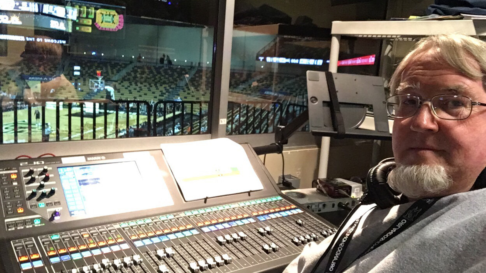 A man seated at an audio console overlooking a basketball court in a live arena.