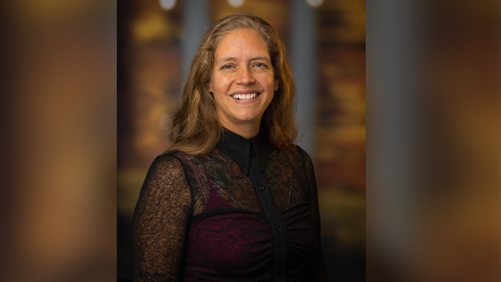 Phoebe Elefante, a woman with shoulder-length light brown hair and a black blouse smiling in front of an out-of-focus brown backdrop.