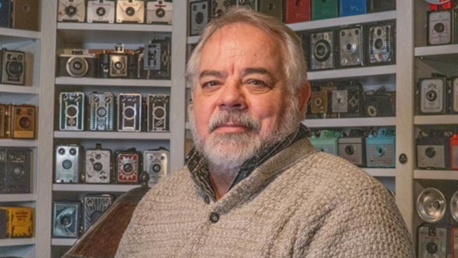 Randy sits in front of a wall covered by shelves of vintage cameras. He is wearing a light brown sweater.