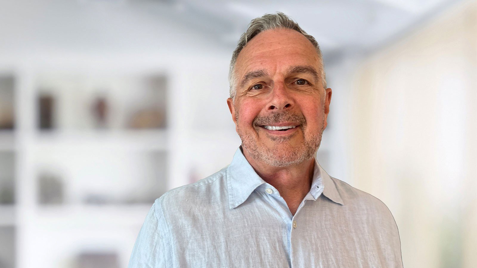 A person wearing a light blue button down while smiling against a white out-of-focus backdrop.