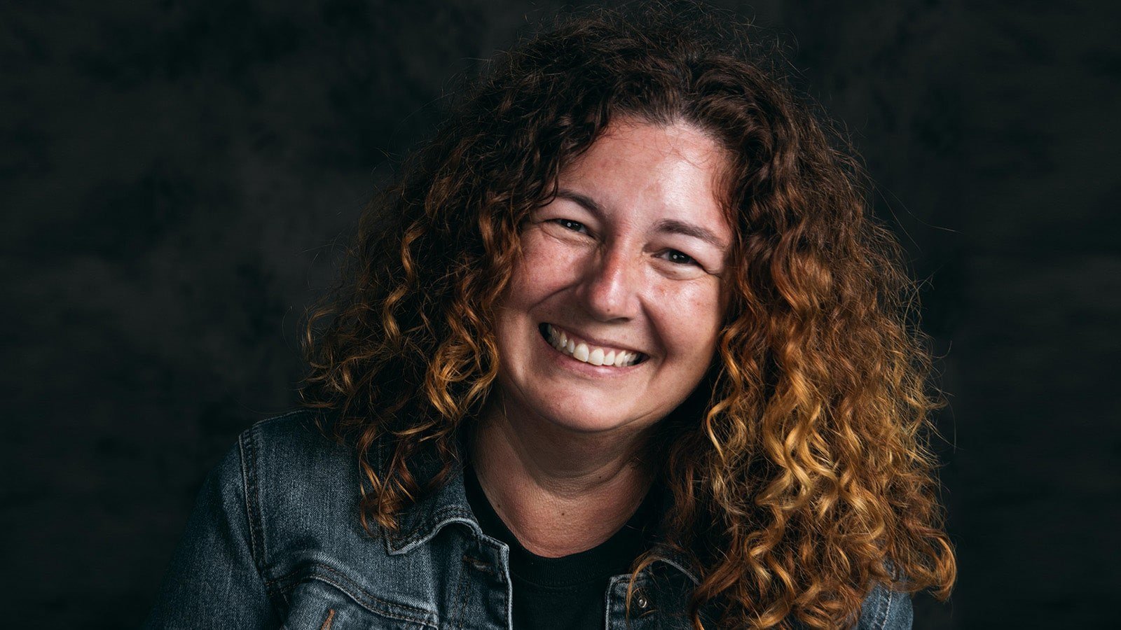 A woman with curly auburn hair smiling while wearing a black tee and denim jacket against a black backdrop.
