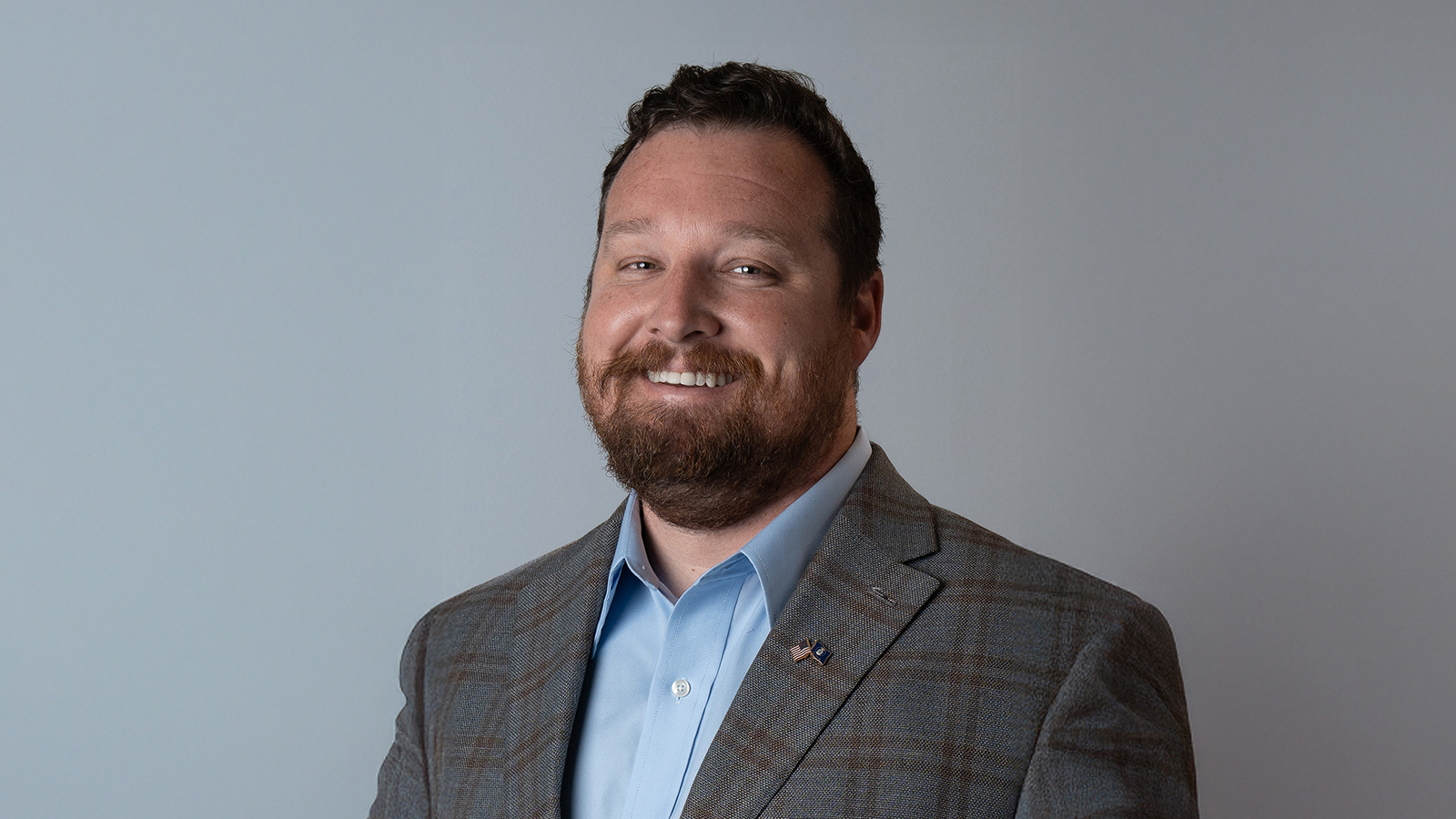 A man with short brown hair and a short brown beard smiles at the camera, wearing a plaid blazer and light blue shirt against a grey background.