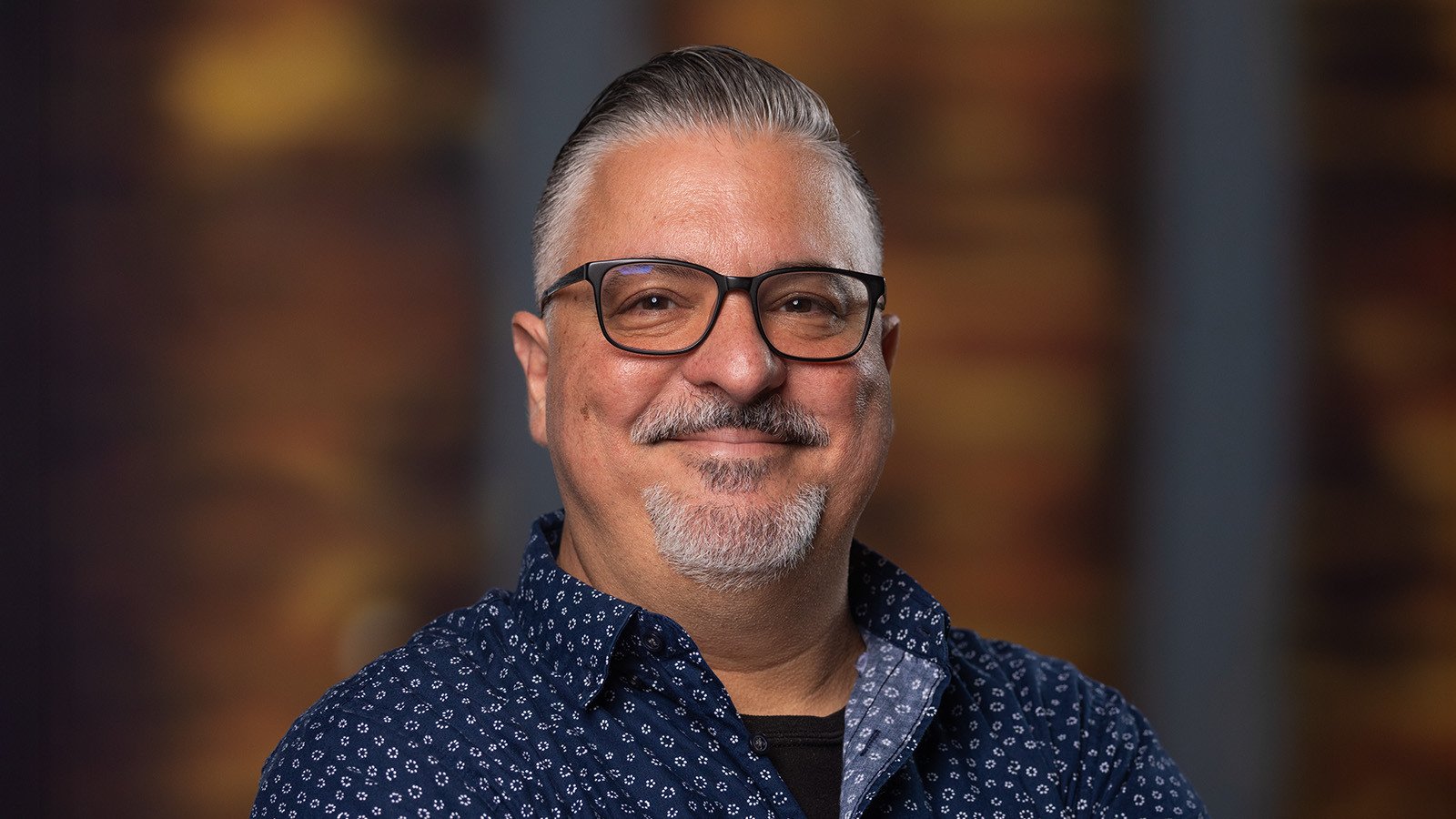 Alex Ginzo smiles at the camera. He is wearing a blue and white collared shirt and glasses, and he has gray hair and a short gray beard.