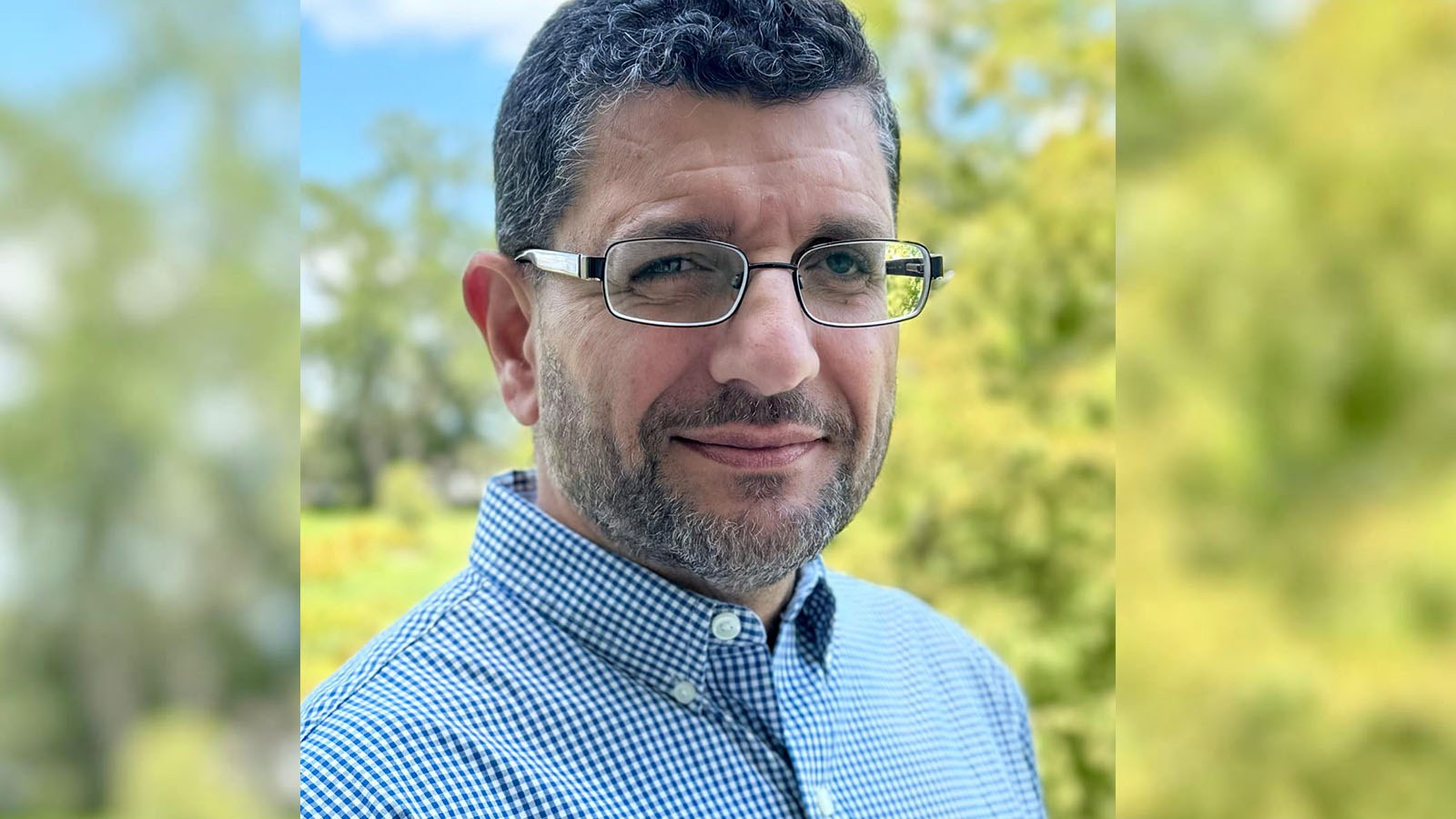 A man with short curly hair, glasses, and a trimmed beard smiles slightly while standing outdoors, the background is out of focus.