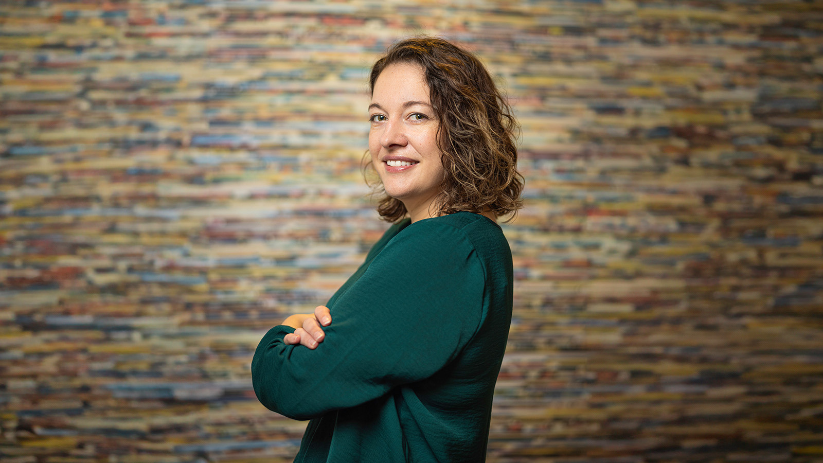 Aron Gelineau stands with her arms crossed and smiles at the camera. She has brown, curly hair and is wearing a teal sweater.