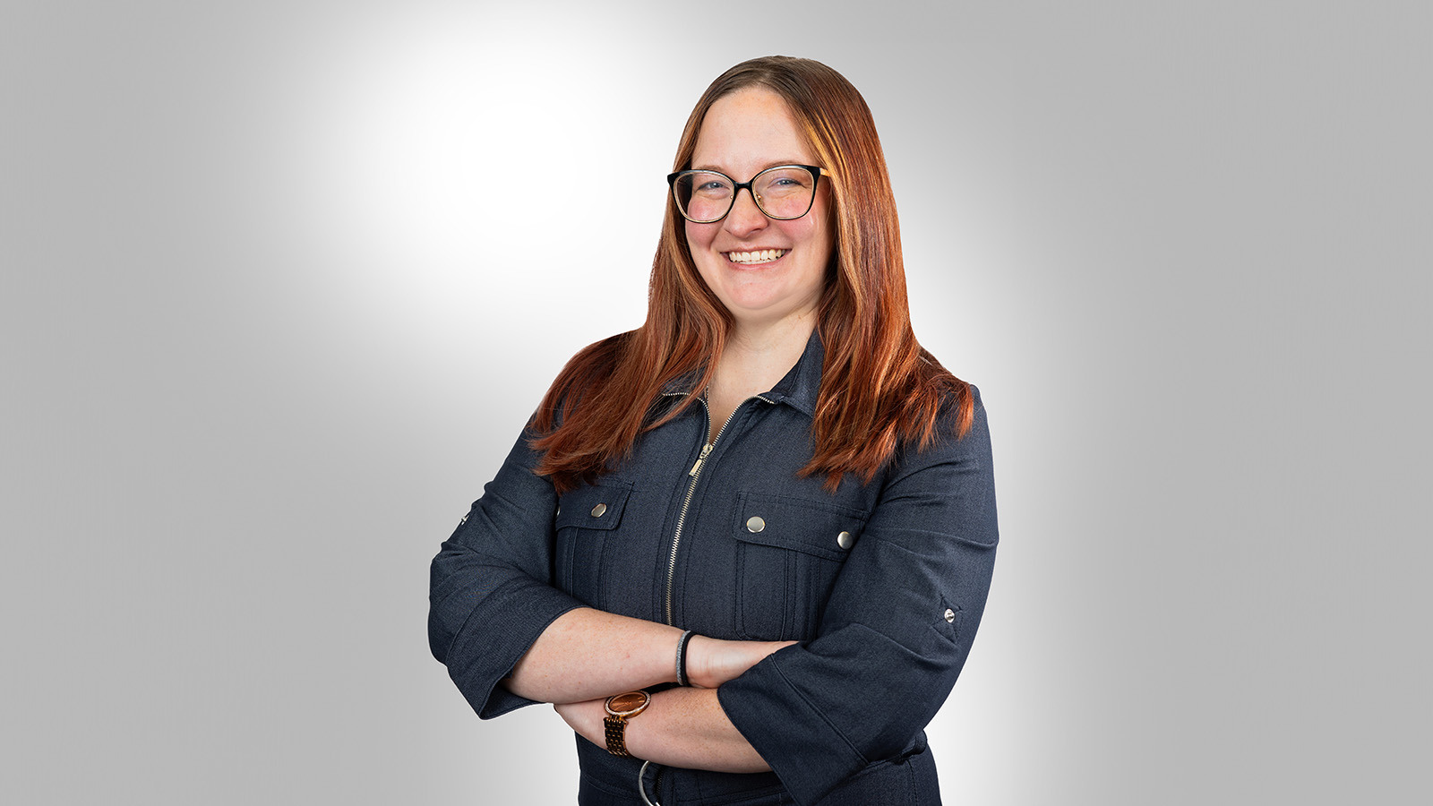 A smiling person with long red hair, glasses, and a blue zip-up jacket, is standing with their arms crossed against a light gradient background.