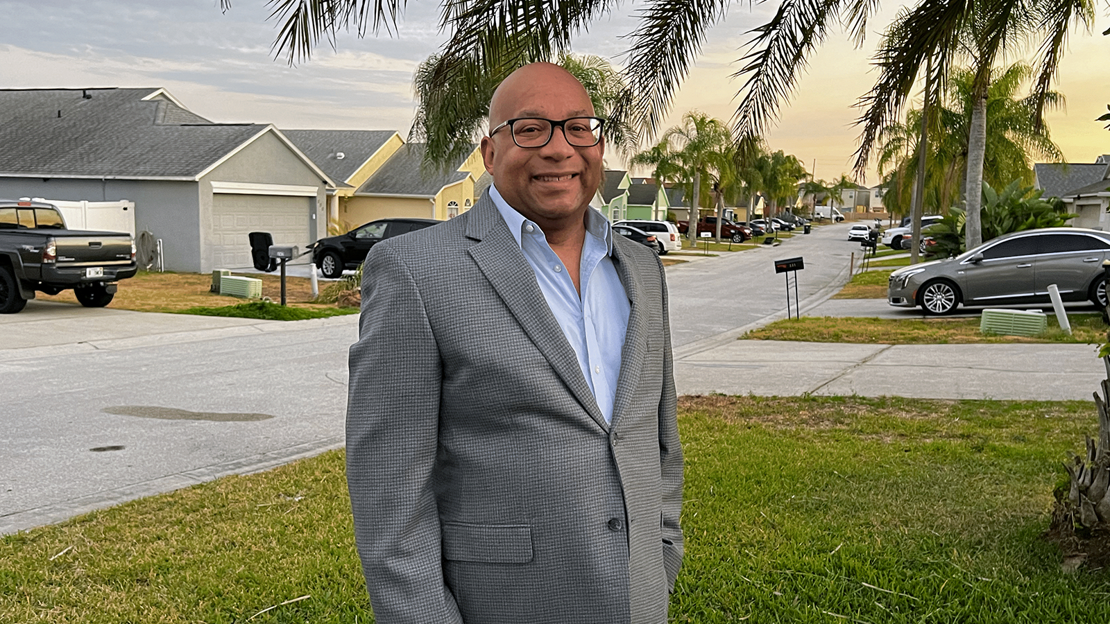 Chris stands outside wearing a grey blazer, blue shirt, and black-rimmed glasses. He is smiling.