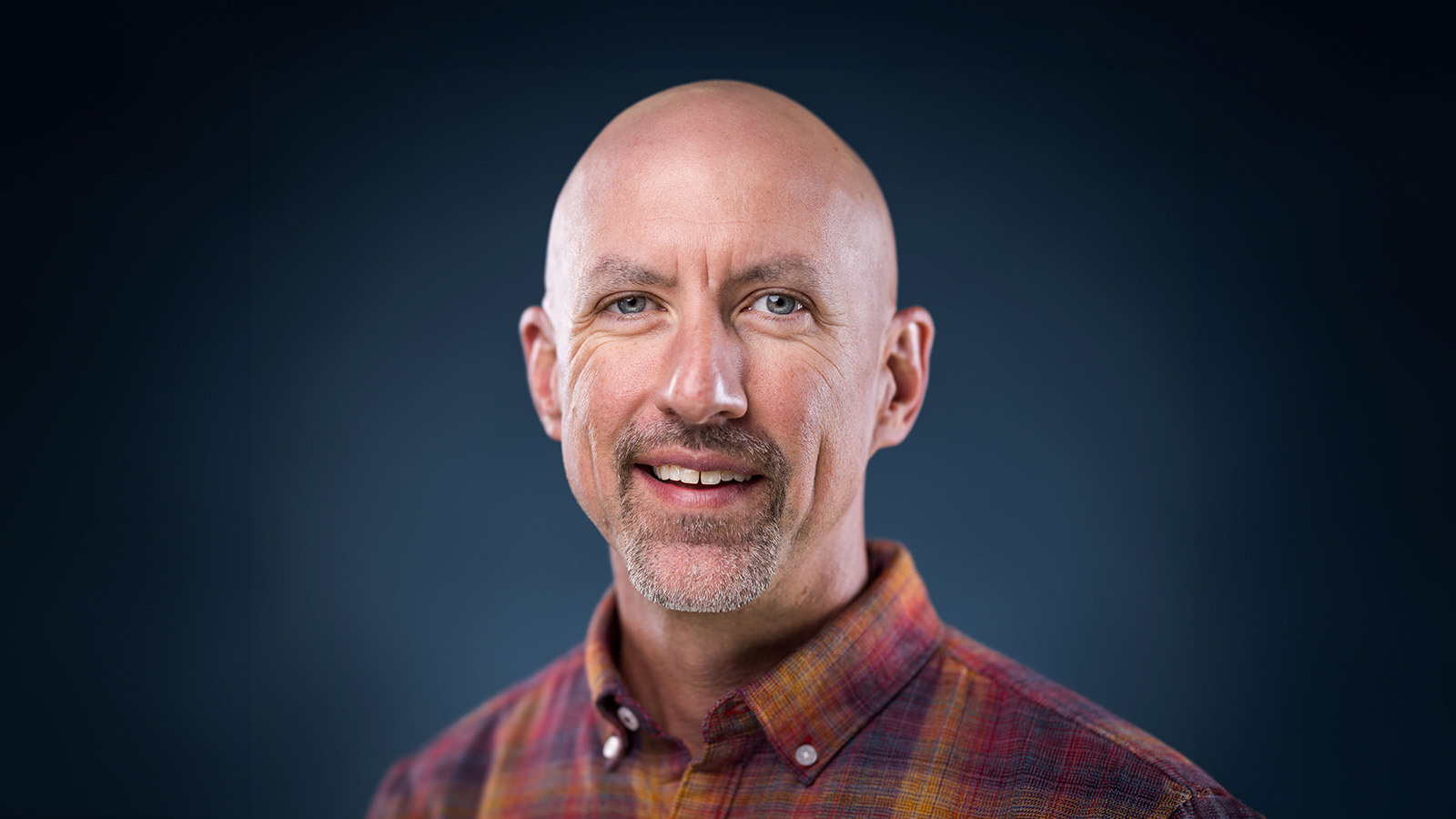 Corbett Compel smiles for the camera in front of a blue background. He is wearing a plaid shirt.