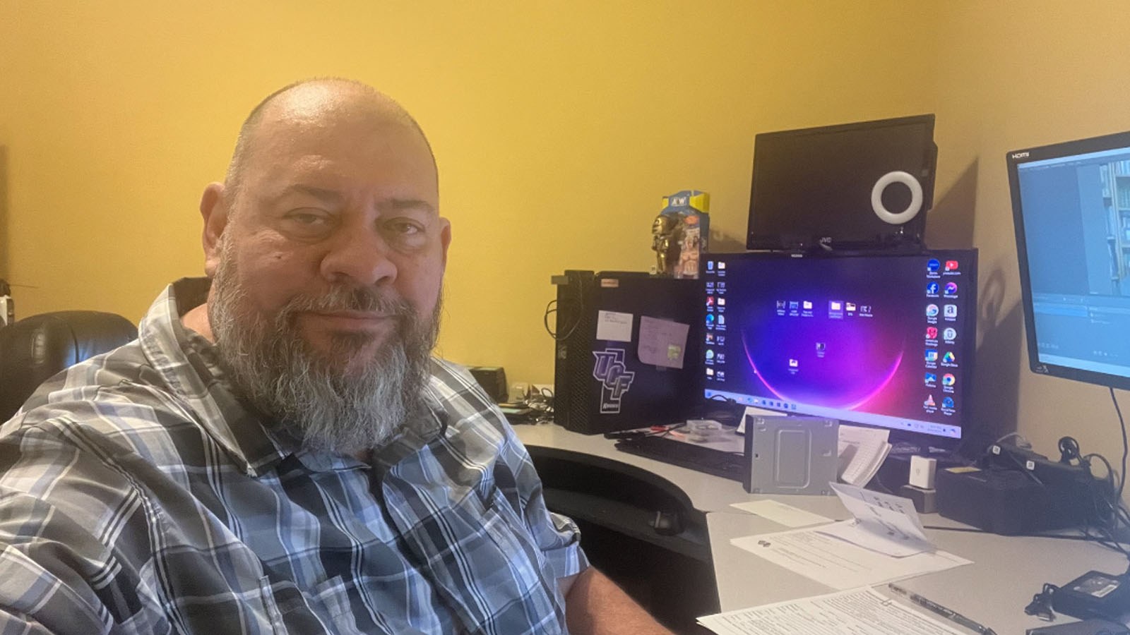 Dan Walker sits in his home office. He has a beard and is wearing a plaid shirt. A desk with several desktop monitors is behind him.