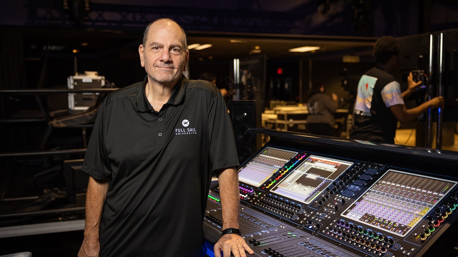 Dave Dean leans against a mixing console in a production space. He is wearing a black Full Sail shirt.