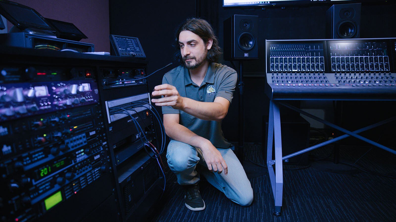 Dave Harris crouches next to broadcast equipment, including mixing consoles and a communications system.