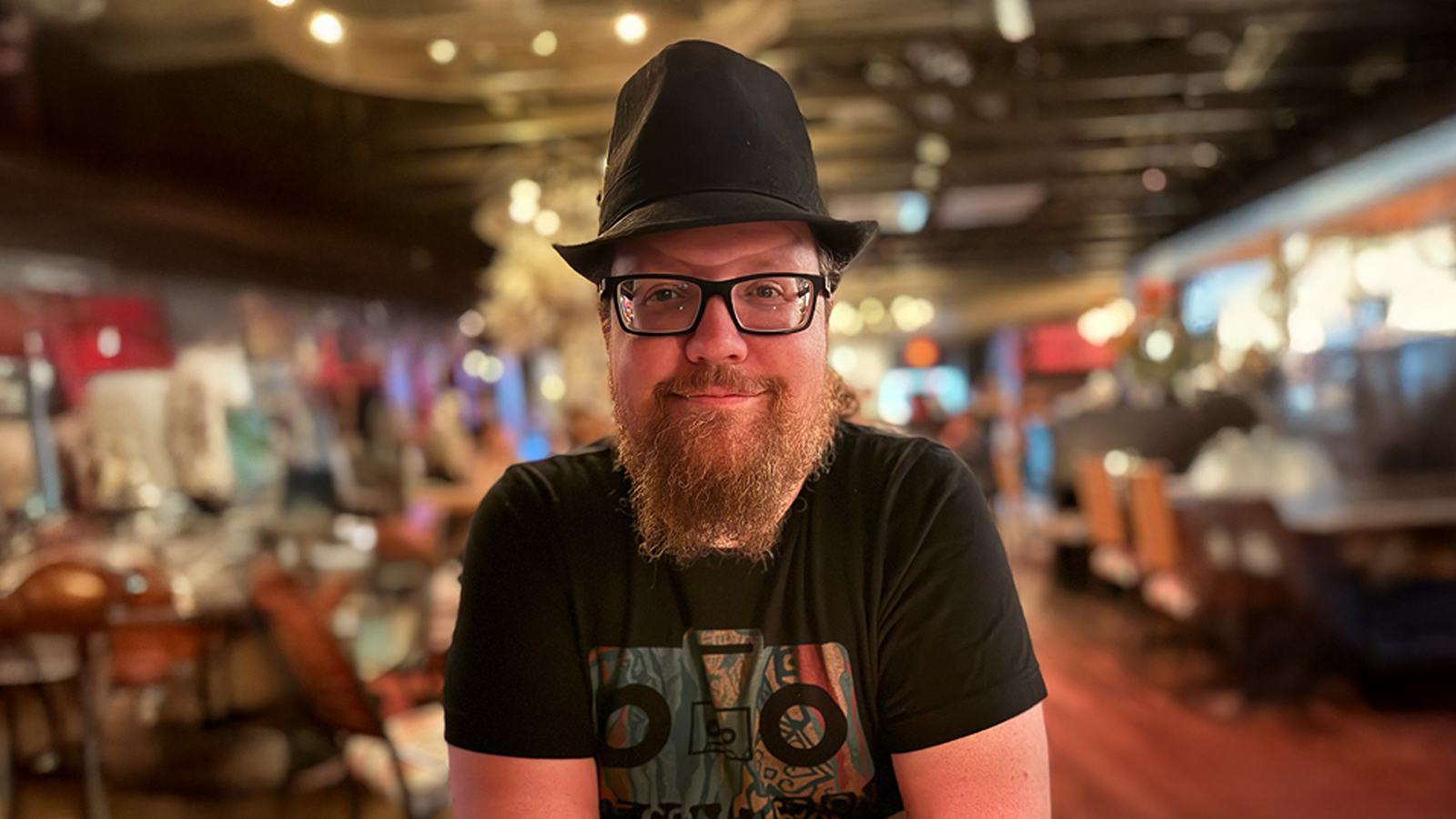 David Dennison sits at a table with his arms crossed. He has glasses and a beard and is wearing a black t-shirt and black fedora.