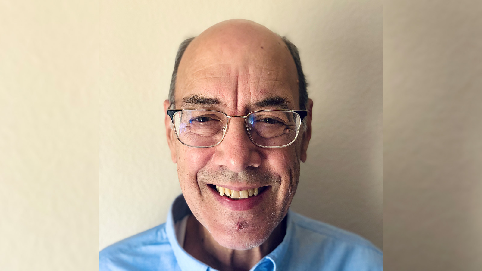 Dr. Carstens stands in front of a white wall wearing a blue shirt and glasses. He is smiling.