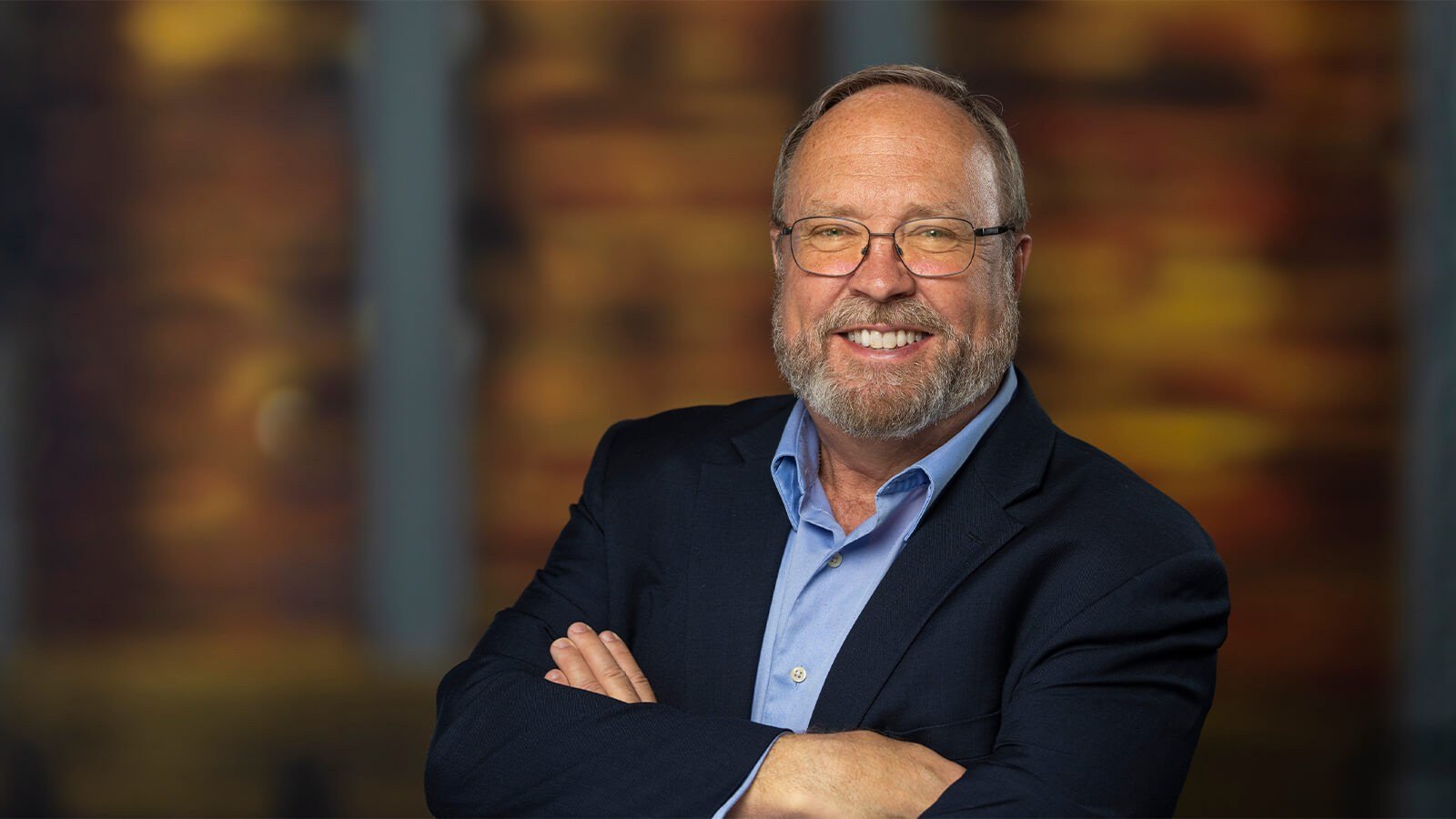 Reo McBride stands with his arms crossed and smiles at the camera. He has a beard and glasses; he’s wearing a blue button-up shirt and a blue blazer.
