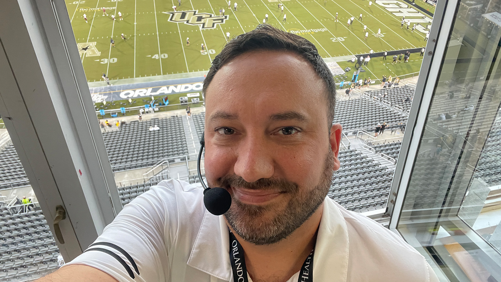 Jeff Sharon wears a headset microphone and smiles at the camera. He is in an announcer’s booth with the UCF football stadium below him.