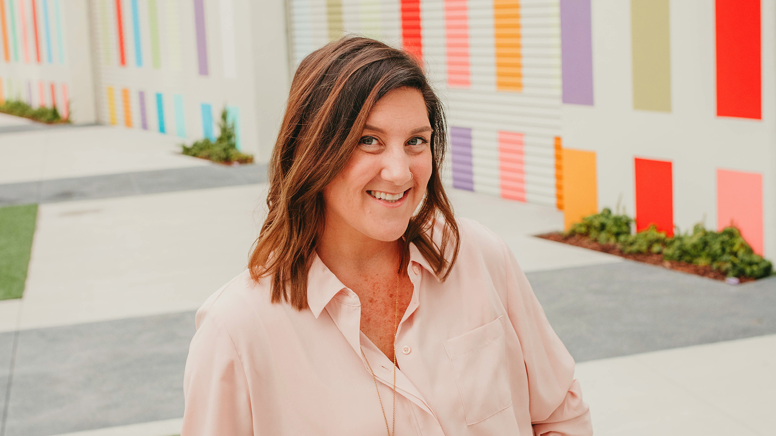 Jenn Miller sits in front of a colorful outdoor wall. She is smiling and wearing a pink shirt and a gold necklace.