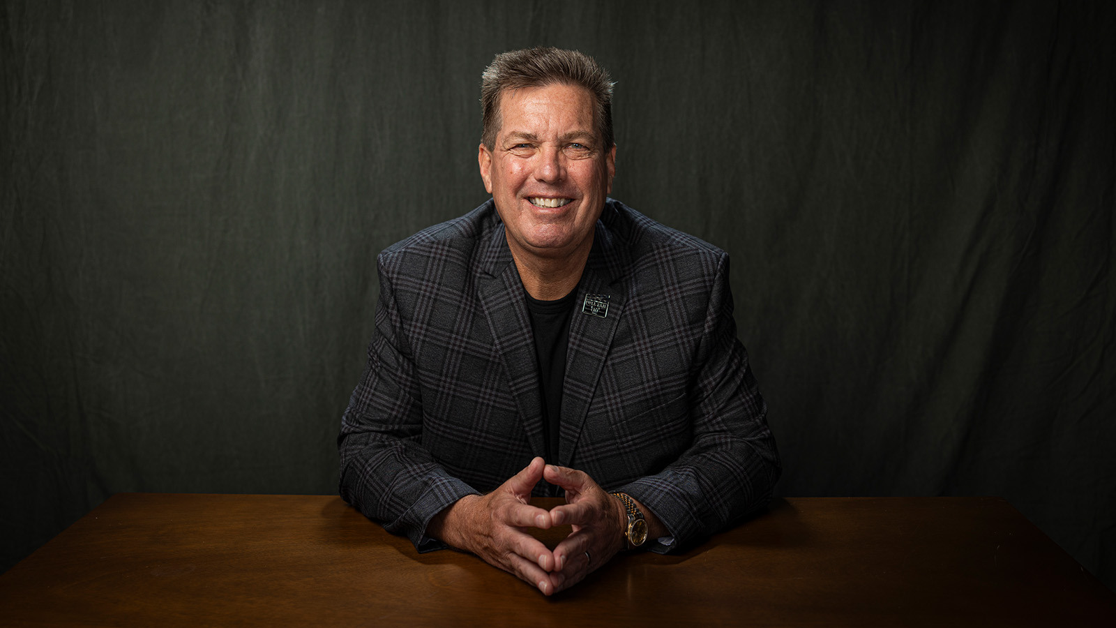 Joe Gornik sits at a wooden table and smiles at the camera. He is wearing a gray plaid jacket with a Full Sail employee pin on the lapel.
